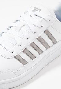 Sneaker in finta pelle bianca con una texture liscia, caratterizzata da strisce verticali grigie, lacci bianchi e suola in gomma.