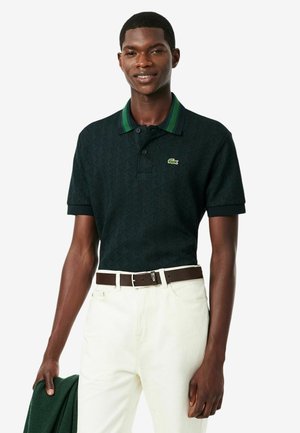 Man draagt een donker groen polohemd met patroon en krokodillenlogo, witte broek en een bruine riem, houdt een groene trui in één hand.