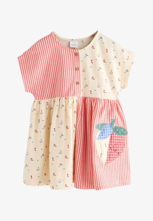 Ung pige iført en kortærmet patchwork kjole med røde striber og blomstermønstre, stående barfodet på en neutral baggrund.