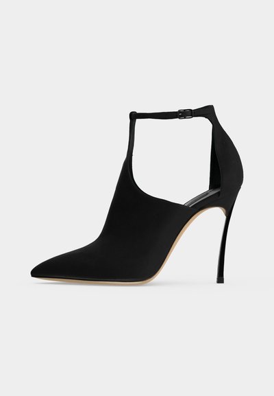 Chaussure à talon haut noire à bout pointu, talon aiguille fin et bride de cheville avec boucle sur fond clair.