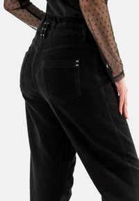 Los pantalones de pana negros presentan una cintura alta, una superficie texturizada, dos bolsillos traseros con detalles de metal y un corte relajado.