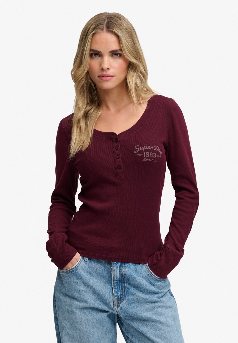 Blonde woman wearing a long-sleeve maroon henley shirt with "SuperDry 1983 Athletics" print and light blue jeans, hands in pockets.