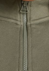 Olivgrön, texturerad tygjacka med en central dragkedja med matt finish och en hög krage.