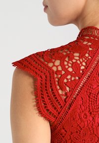 Röd spetsblus med intrikata mönster, texturerad broderi och utsmyckad axeldesign. Nära bild fångar detaljer och hantverk.