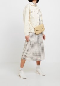 Krémová džínová bunda přes kostkované šaty s vrstvenou sukní. Béžová kabelka přes rameno a bílé špičaté kotníkové boty doplňují vzhled.