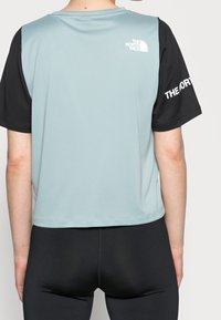 Osoba stojí zády, má na sobě světle tyrkysový a černý sportovní top s logem "The North Face", doplněný černými legínami.