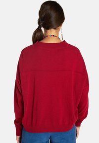 Roter Pullover mit lockerer Passform, geripptem Halsausschnitt und überschnittenen Schultern. Der Stoff wirkt weich und hat leicht strukturierte Details.