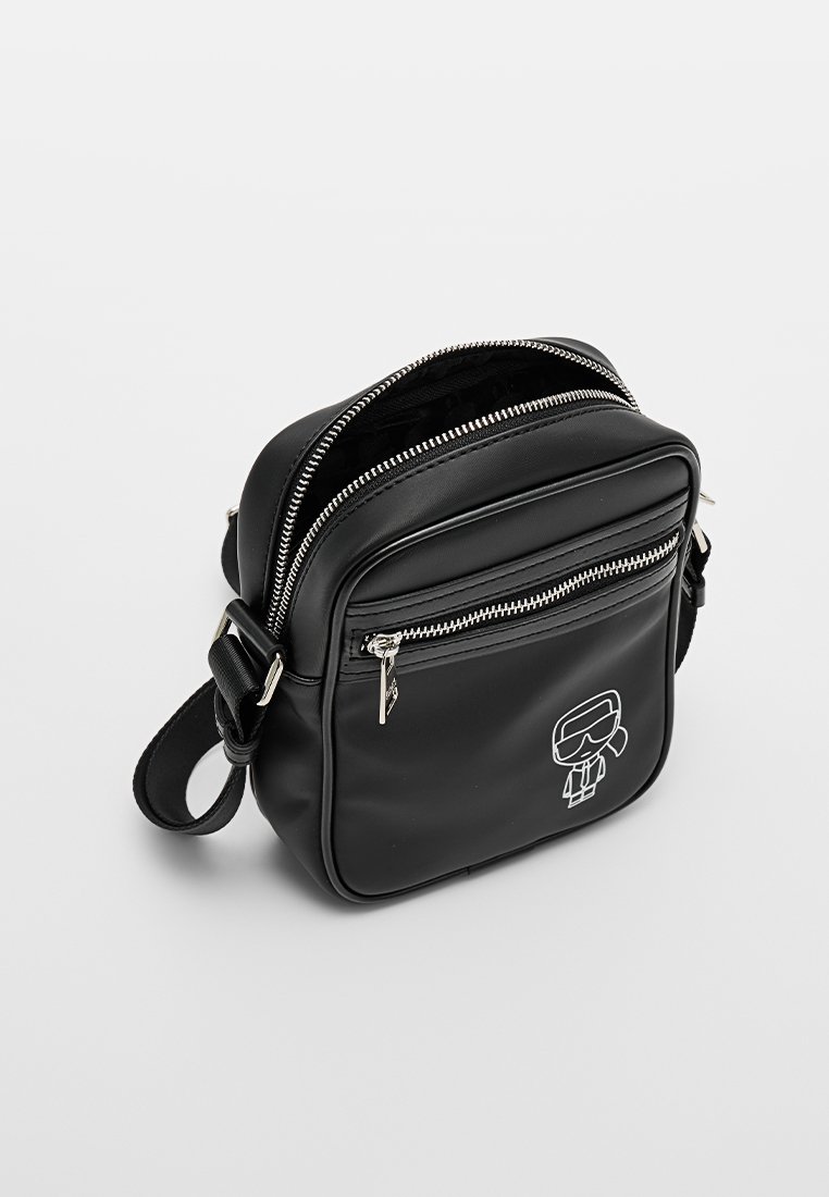 Bolso bandolera de cuero sintético negro con cierre de cremallera, bolsillo frontal y una ilustración gráfica en la parte delantera. Detalles en hardware plateado.