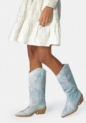 Frau, die kniehohe blaue Schlangenlederstiefel und ein weißes Spitzenkleid trägt, steht mit übereinander geschlagenen Beinen.