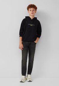 Zwarte hoodie met een grafische afbeelding aan de voorkant, gecombineerd met donkere jeans en lichte sneakers. Model staat ontspannen met de handen in de zakken, op een witte achtergrond.