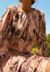 Blomsterkjole i blødt pink stof med lange, flagrende ærmer. Har mørk lilla og grønne blomsterpattern, samlet halsudskæring og midi-længde nederdel.