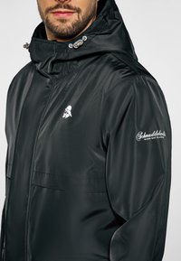 Svart vattentät jacka med hög krage, justerbara dragsnören och en vit logotyp på bröstet. Slät, elegant textur med en minimalistisk design.