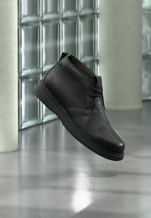 Schwarzer Leder-Knöchelstiefel mit Schnürsenkeln, schwebend über einem polierten grauen Boden nahe einer Glassteinwand und weißen Säulen.