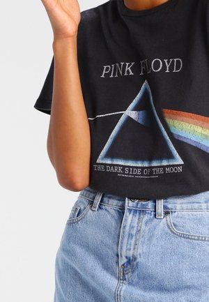 Personne portant un t-shirt noir Pink Floyd avec le prisme et le design arc-en-ciel de Dark Side of the Moon, ainsi qu'un jean en denim bleu clair.