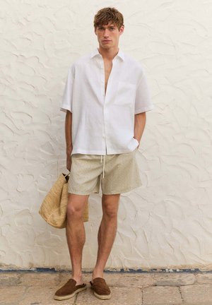 Camicia bianca di lino con taschino, pantaloni corti beige a fantasia e espadrillas marroni. Tieni in mano una borsa di paglia intrecciata, in piedi su una superficie in pietra testurizzata.