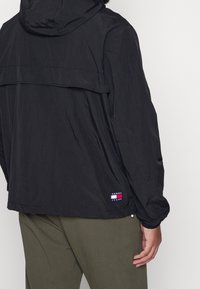 Lekka czarna kurtka z kapturem, o luźnym kroju, ze ściągaczami na rękawach i rozcięciem z tyłu. Na dole przy naszyciu znajduje się naszywka z logo w kolorach czerwonym, białym i niebieskim.