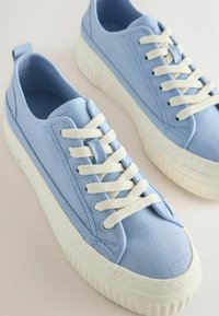Paire de baskets en toile bleu clair avec des semelles épaisses en caoutchouc blanc et des lacets blancs, vues d’un angle supérieur sur une surface blanche.