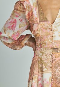 Sukienka w kwiatowy wzór z miękkiego materiału w odcieniach różu, beżu i złota. Posiada falbaniaste rękawy oraz dekoracyjne guziki na froncie.