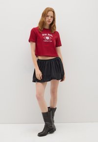 Top corto de algodón rojo con estampado gráfico, combinado con una falda negra con fruncidos. La modelo lleva botas de cuero negro hasta la rodilla. Atuendo sencillo y casual.