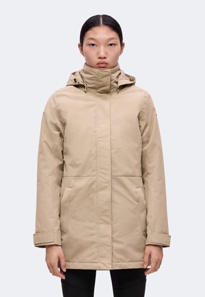 Frau trägt eine beige, gefütterte Winterjacke mit Kapuze und Fronttaschen, steht frontal vor einem schlichten Hintergrund.