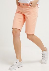 Pantalones cortos de mezclilla melocotón con un ajuste entallado, que cuentan con bolsillos frontales y un cierre de botón. Combinados con zapatillas grises.