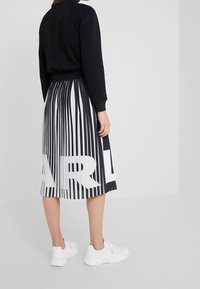 Schwarzes Langarmoberteil kombiniert mit einem plissierten schwarz-weißen Rock mit vertikalen Streifen und übergroßem Textprint. Dazu weiße Sneaker getragen.