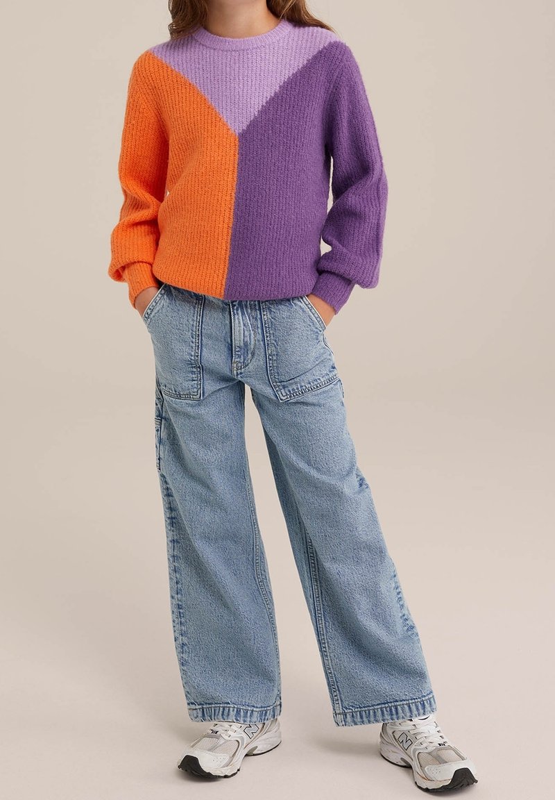 Personne portant un pull colorblock en tricot orange, violet et lavande avec un jean à jambes larges bleu et des baskets blanches, mains dans les poches.