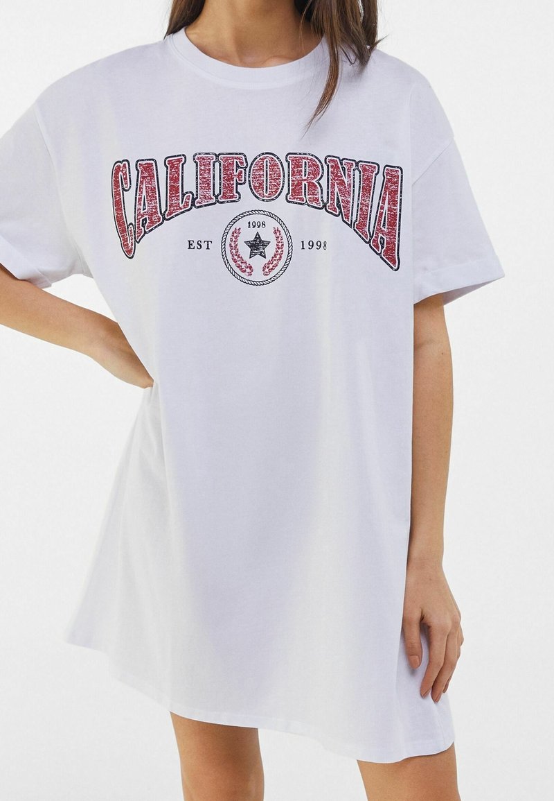Femme portant une robe T-shirt blanche surdimensionnée avec le texte rouge « CALIFORNIA » et un emblème étoile noire, main sur la hanche, sur fond blanc uni.