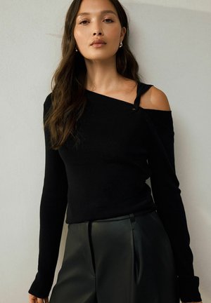 Frau mit langen dunklen Haaren, die ein schwarzes asymmetrisches Oberteil mit kalten Schultern und schwarze hochtaillierte Hosen trägt, steht vor einer hellgrauen Wand.