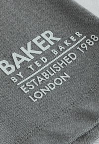 Grijze stof met het reliëf van witte tekst waarop staat "BAKER by TED BAKER ESTABLISHED 1988 LONDON," met een getextureerd oppervlak en gestikte randen.