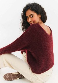Bordowy dzianinowy sweter o luźnym kroju, szerokich rękawach i dekolcie w kształcie litery V z tyłu. Miękka faktura połączona z beżowymi spodniami i brązowymi butami.