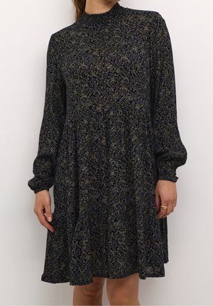 Kvinde iført en sort kjole med lange ærmer og høj hals, dekoreret med et lille blomsterprint i afdæmpede blå og beige nuancer.