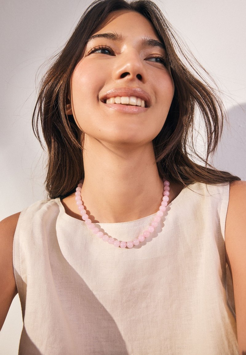 Pink bead necklace on a bare neck, paired with an off-white, sleeveless linen top. Soft lighting highlights the texture and colors.