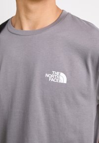 Szara bawełniana koszulka z okrągłym dekoltem, z białym logo przedstawiającym wzór góry na lewej stronie klatki piersiowej. Miękka faktura, standardowy krój.