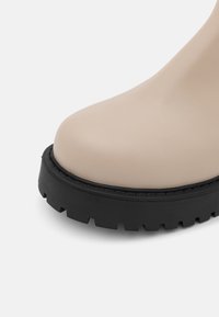Bežová kožená kotníková bota s kulatou špičkou, která má silnou černou gumovou podrážku s hlubokým vzorem pro lepší trakci.
