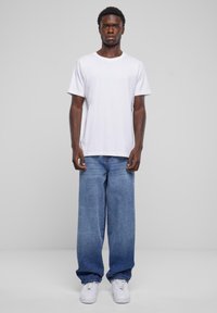 Vit bomull t-shirt med kort ärm, i kombination med baggy blå denimjeans. Modellen bär vita sneakers och står mot en grå bakgrund.