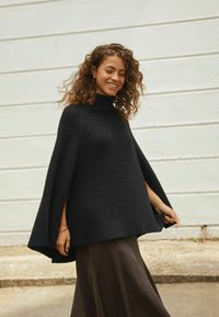 Woman with curly hair wearing a black knit poncho and brown skirt, smiling with eyes closed, standing outdoors near a white wall.