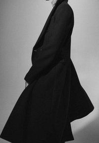Homme portant un long manteau noir à ourlet fluide, posant devant un fond clair uni, cadré sous les épaules.