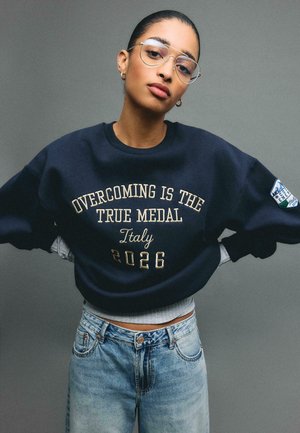Femme aux cheveux gominés en arrière, portant des lunettes, un sweat-shirt marine avec l'inscription « Surmonter est la vraie médaille Italie 2026 » et un jean bleu clair.