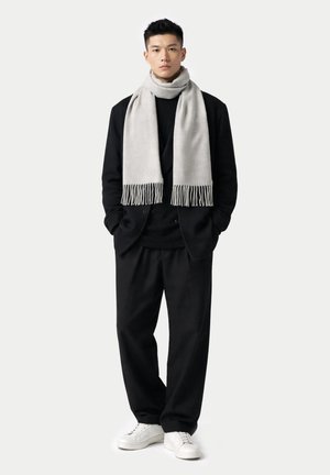 Mann steht mit den Händen in den Taschen, trägt schwarzen Anzug, schwarzes Hemd, weiße Turnschuhe und einen hellgrauen Schal mit Fransen.