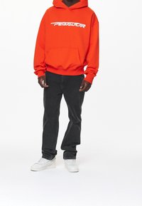 Leuchtend orangefarbener Hoodie mit weißem Logo, Fronttasche, gerippten Bündchen, kombiniert mit schwarzer Jeans und weißen Turnschuhen. Einfaches, lässiges Design.