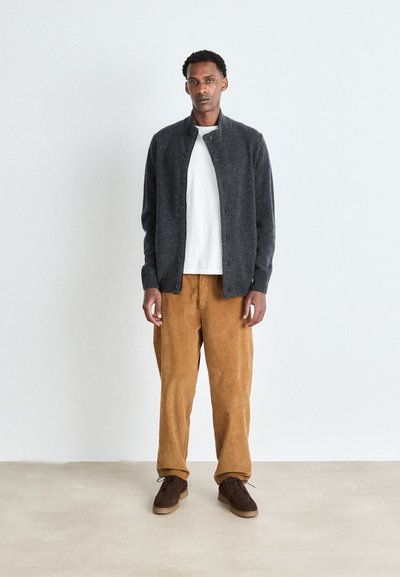 Cardigan gris foncé en tricot texturé, t-shirt blanc et pantalon en velours côtelé beige. Des chaussures marron complètent la tenue. Se tenant devant un fond clair.