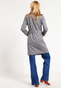 Abrigo de lana gris con cintura ceñida, de longitud media a los muslos y mangas largas. Combinado con jeans de pierna ancha azules y zapatos de tacón rojo.