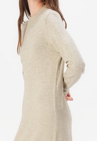 Femme portant une robe en maille beige à manches longues, se tenant avec une main derrière le dos contre un fond blanc uni.