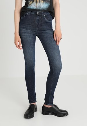 Henkilö pukeutuneena tummansinisiin kapealahkeisiin farkkuihin, mustiin nauhallisiin kenkiin ja mustaan kuvalliseen t-paitaan, seisomassa yksiväristä vaaleaa taustaa vasten.