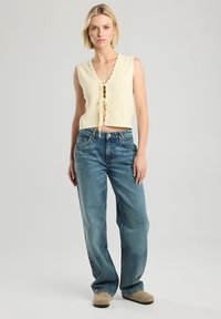 Cream knitted crop top with scalloped edges and tie front detail, paired with light blue wide-leg jeans and beige slip-on shoes.