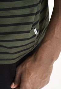 Grønstribet t-shirt med sorte striber, blødt stof, og et lille mærke på siden nær sømmen. Modellen holder stoffet med den ene hånd.
