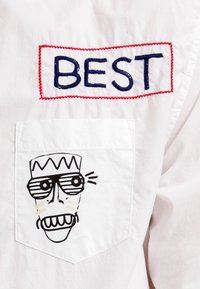 Vit skjorta med röd och blå broderad "BÄST"-etikett. Framtikett har ett tecknat ansikte i svart och vitt med solglasögon och en krona.