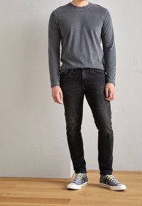 Grå långärmad skjorta, svarta jeans med slitage och grå sneakers med gröna snören, mot en enkel, ljus bakgrund.