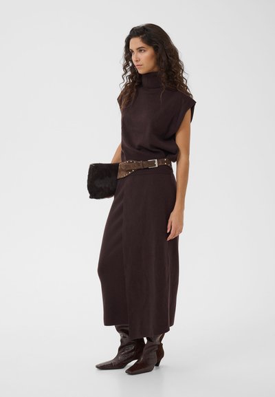 Femme aux longs cheveux bouclés portant une robe sans manches à col roulé marron foncé, une large ceinture, des bottes assorties et tenant une pochette en fourrure.